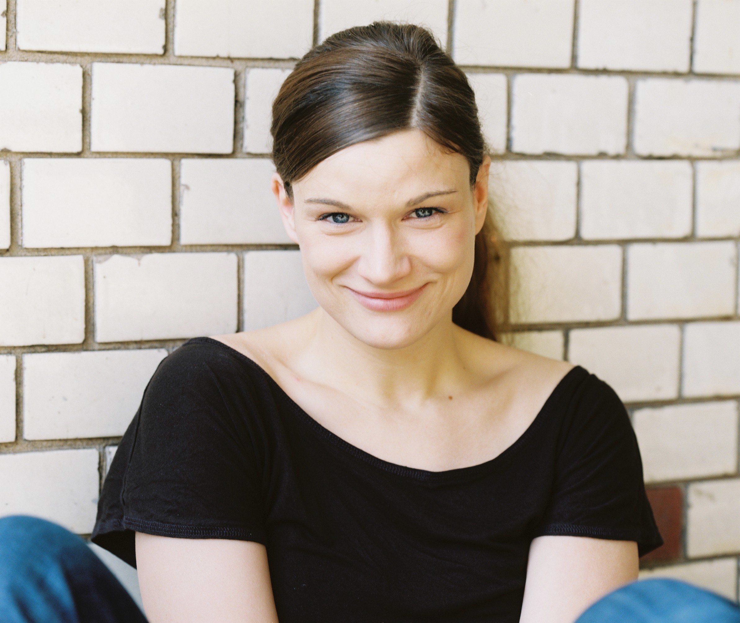 Das Foto zeigt Sandra Rasch. Sie hockt auf dem Boden vor einer weißen Mauer und lächelt. Sie hat braune Haare in einem Zopf und trägt ein schwarzes T-Shirt. 